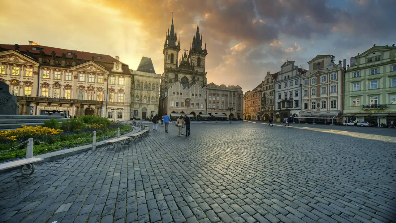 Wide view of Prague’s Old Town Square at sunset, featuring cobblestone streets, colorful historic buildings, and the twin Gothic spires of the Church of Our Lady before Týn with people walking through the square. 