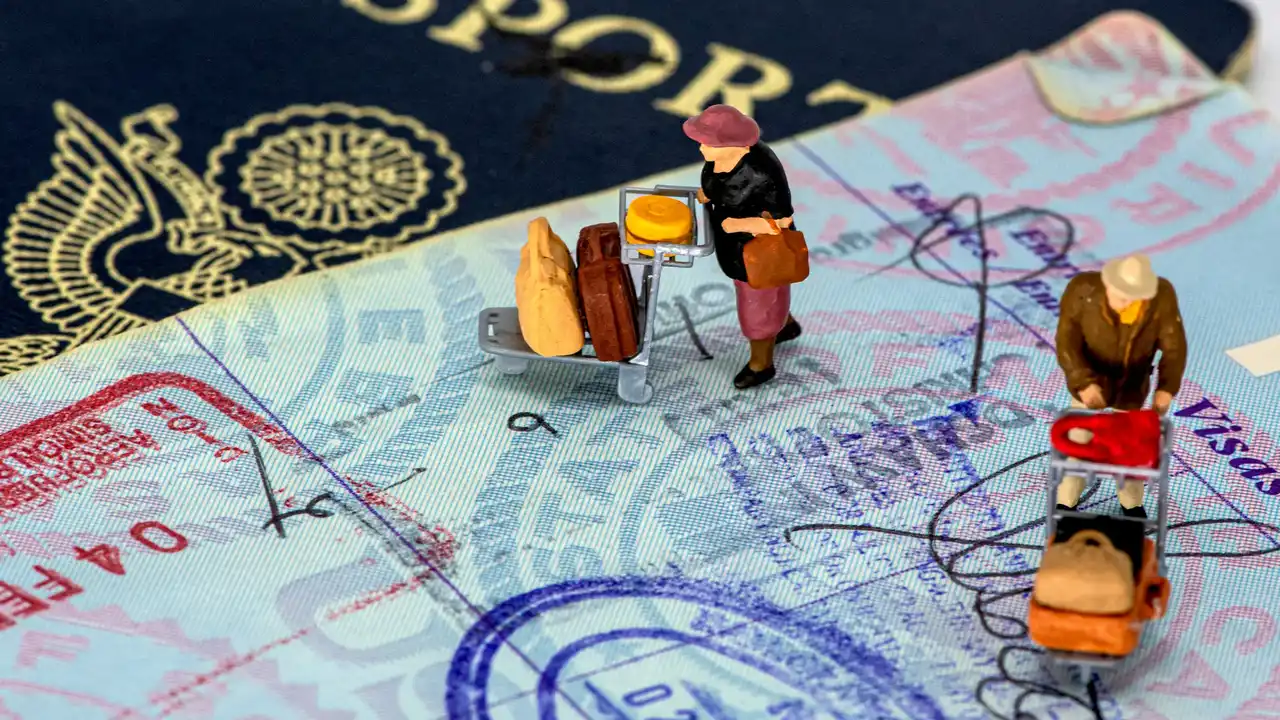 A close-up view of miniature traveler figurines with luggage carts standing on a passport page filled with colorful entry and exit stamps. Behind the visa page, a dark blue passport cover with a gold embossed seal is partially visible.
