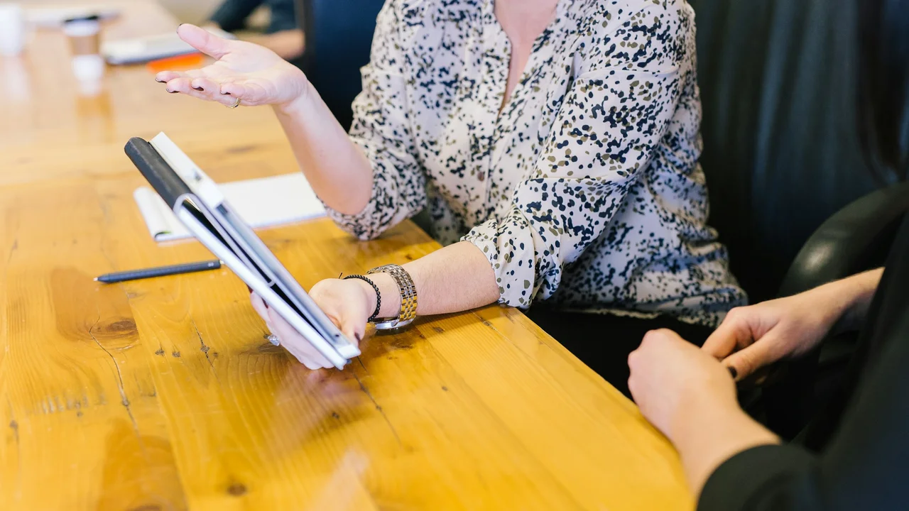 An overhead shot of two people seated at a long, light-colored wooden table during a meeting. One person, wearing a patterned cream and black blouse, holds a white tablet and gestures with their other hand while speaking. Only the hands and torso of the second person are visible on the right. A stylus and a notebook are also on the table.