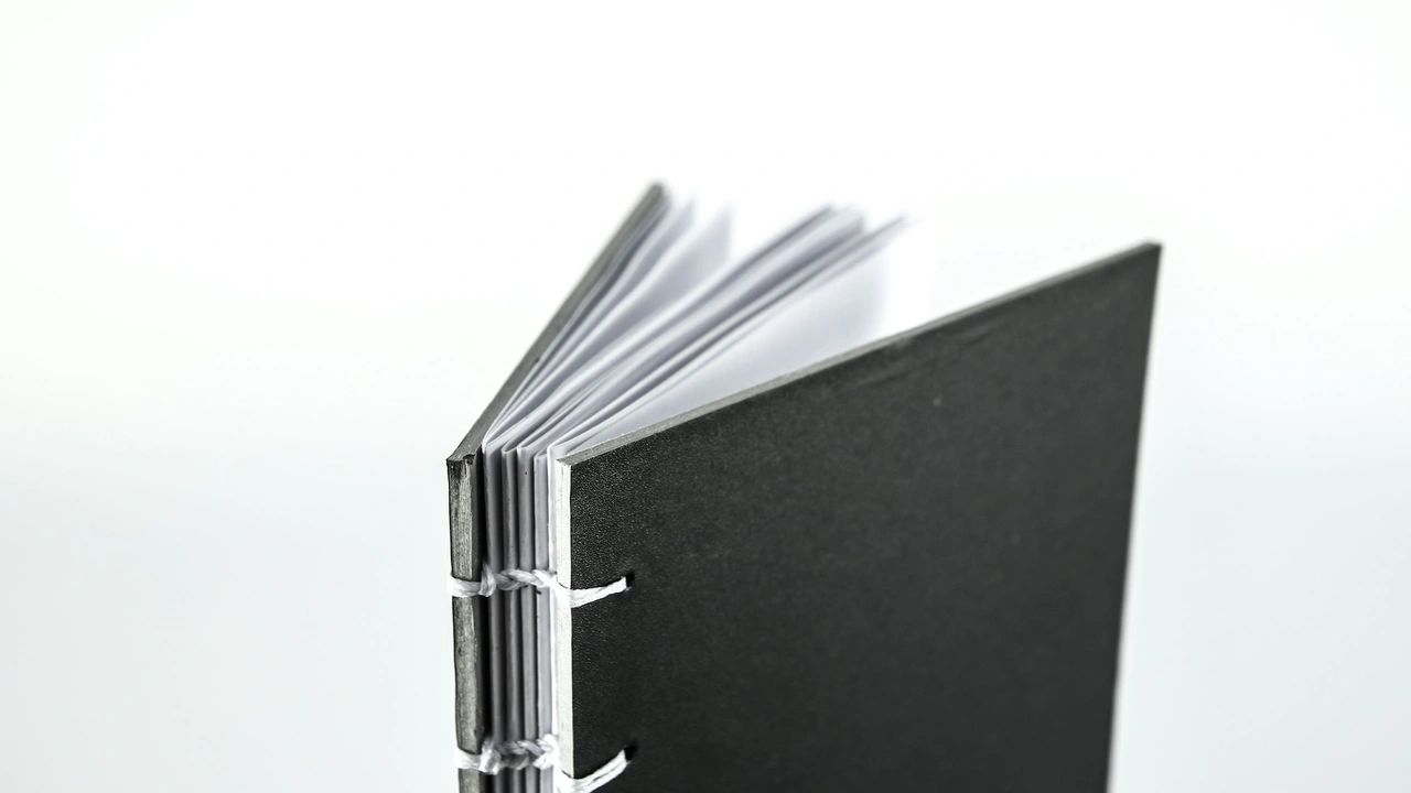 A close-up, minimalist shot of a small, hand-bound booklet with a thick black cardstock cover. The booklet is slightly open, showing several white interior pages. The binding is visible on the spine, featuring white thread stitched through the cover and pages in a clean, functional style against a stark white background.