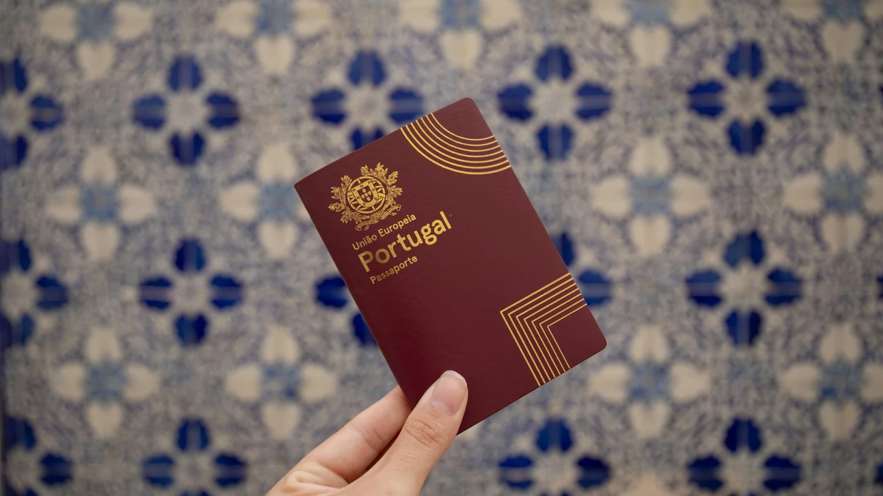 A person's hand holds a burgundy Portuguese passport upright against a blurred background of traditional blue and white azulejo tiles. The passport cover features the gold national coat of arms and the words