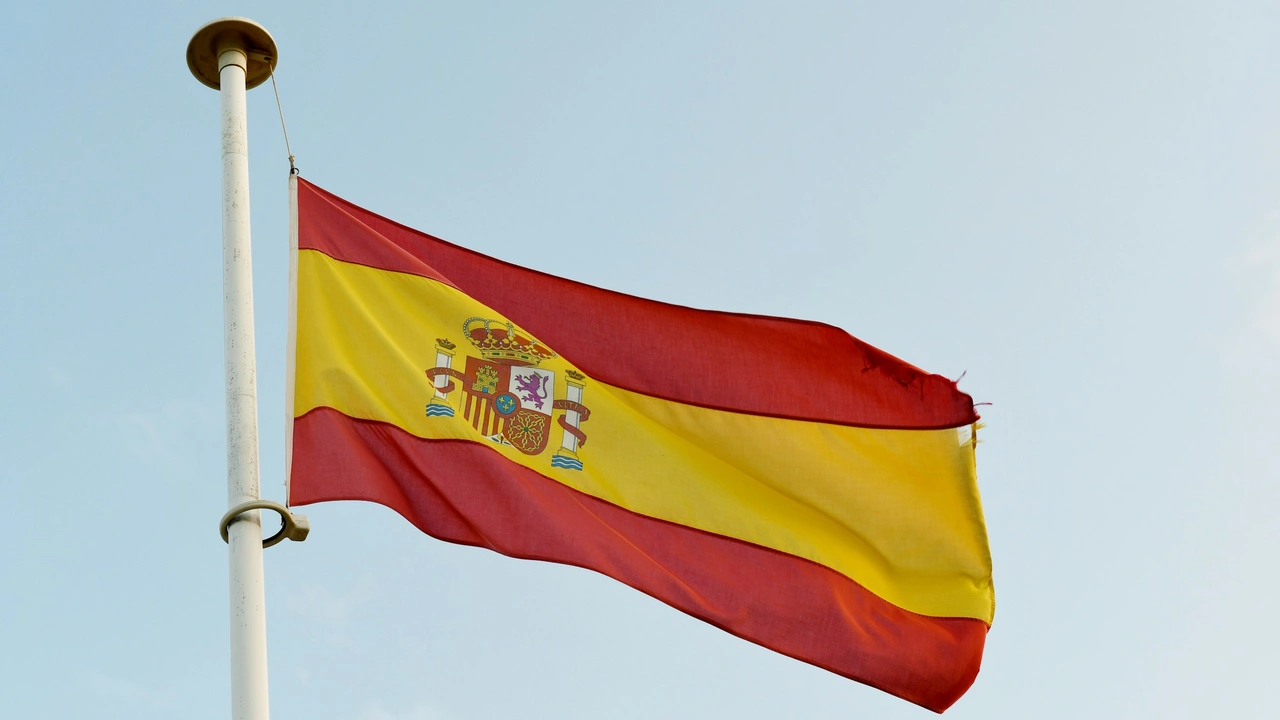 he national flag of Spain, featuring three horizontal stripes—red, yellow (double width), and red—with the Spanish royal coat of arms positioned toward the left of the yellow band. The flag is attached to a white flagpole and waves in the wind against a pale, clear sky.