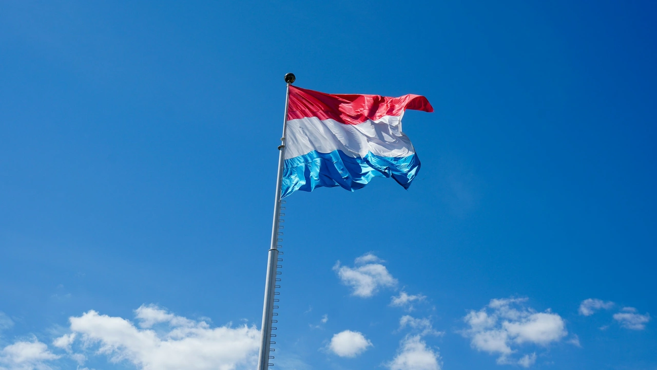 he national flag of Luxembourg, featuring horizontal stripes of red, white, and light blue, flies from a tall silver flagpole against a clear blue sky with sparse, wispy white clouds