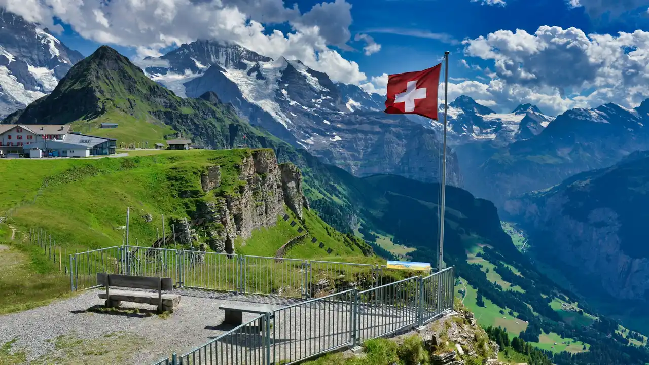 A panoramic view of the Swiss Alps under a bright blue sky with scattered clouds. In the foreground, a gray metal railing encloses a lookout point featuring two wooden benches. To the right, the red and white flag of Switzerland waves from a tall flagpole. The background showcases lush green slopes, rugged rocky cliffs, and towering snow-capped mountain peaks.