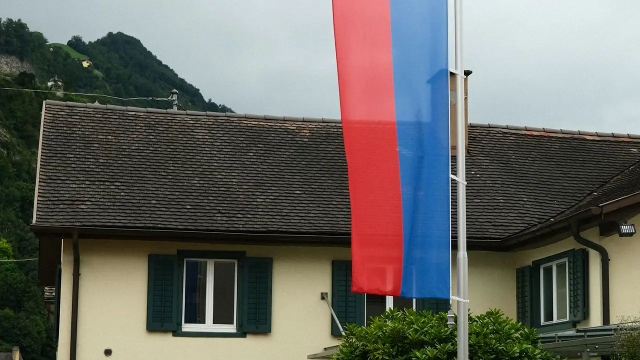 A bright outdoor photograph of the national flag of Liechtenstein waving from a tall silver flagpole against a clear blue sky. The flag features two horizontal bands of blue and red with a gold princely crown in the upper hoist corner. In the background, the modern, light-colored Liechtenstein Parliament building (Landtag) in Vaduz is visible, partially framed by the bare branches of trees.